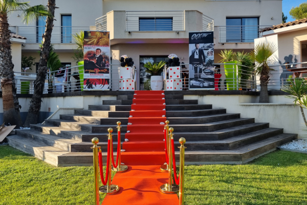 une maison avec une décoration chic avec un tapis rouge le long de l'escalier, des poteaux dorés le long du chemin. En haut de l'escalier se trouve des affiches de films James Bond, des dés et des cartes.