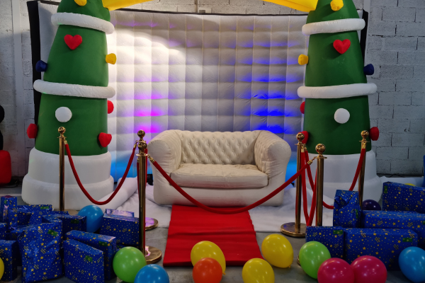 un fauteuil blanc chesterfield est entouré d'une décoration de noël (arche gonflable en sapin, fausse neige, cadeaux sur les côtés et ballons). Avec des poteaux dorés et tapis rouge qui habillent élégamment le fauteuil.